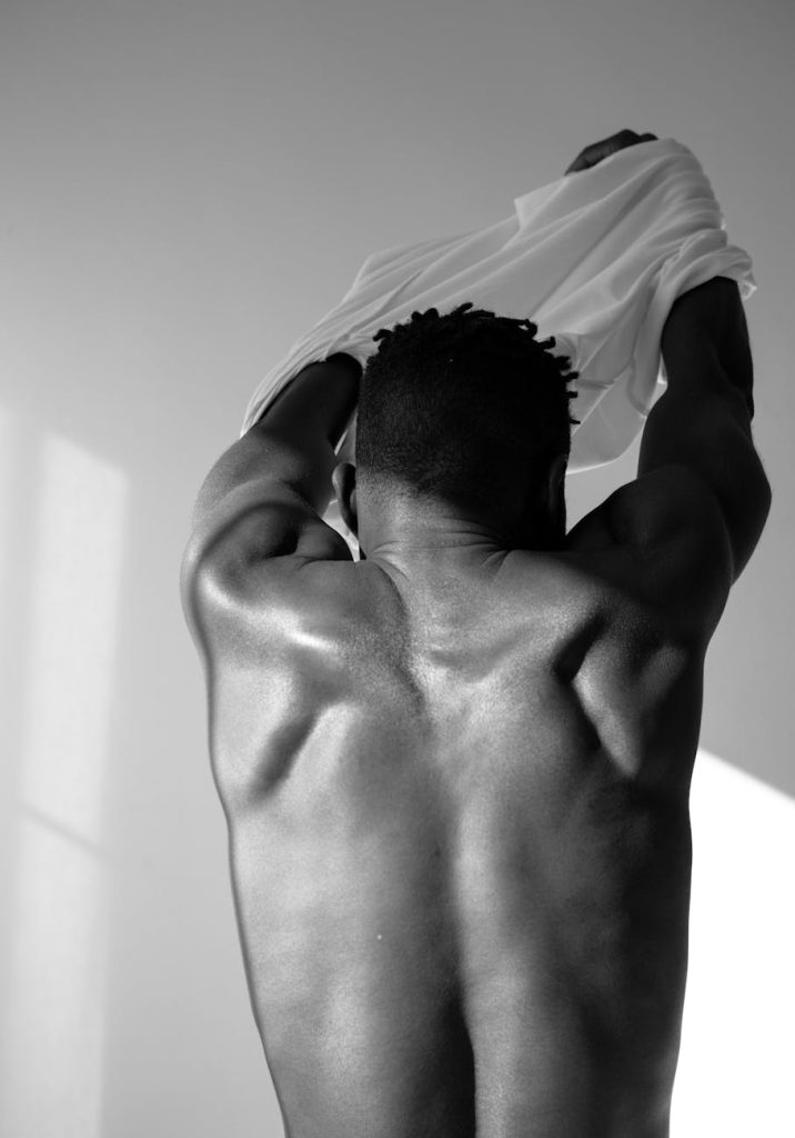 Grayscale image of a shirtless muscular man from behind removing his shirt.