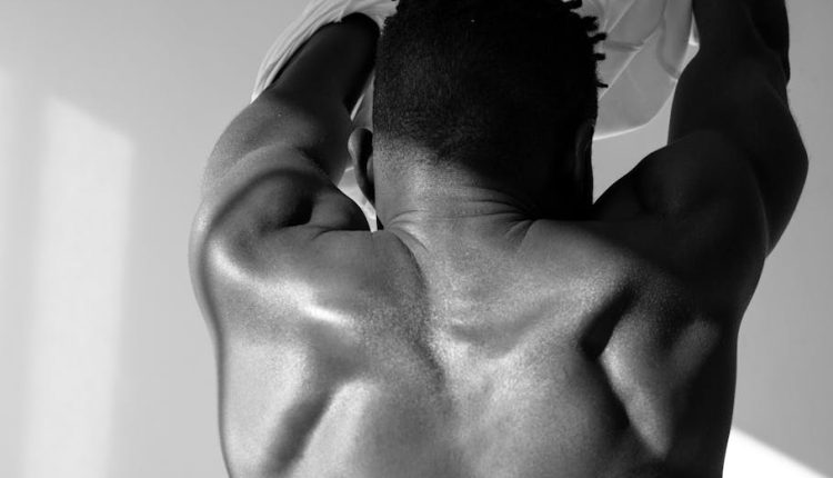 Grayscale image of a shirtless muscular man from behind removing his shirt.