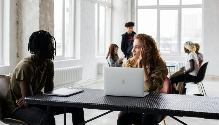 Concentrated young multiethnic coworkers in casual clothes sitting at table with laptops and discussing project details in light loft workspace